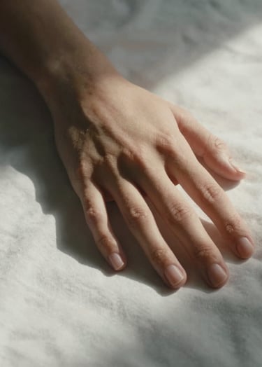 Minimalist close-up of human hands gently resting on soft white fabric, lit with a warm, cinematic glow and deep charcoal shadows.