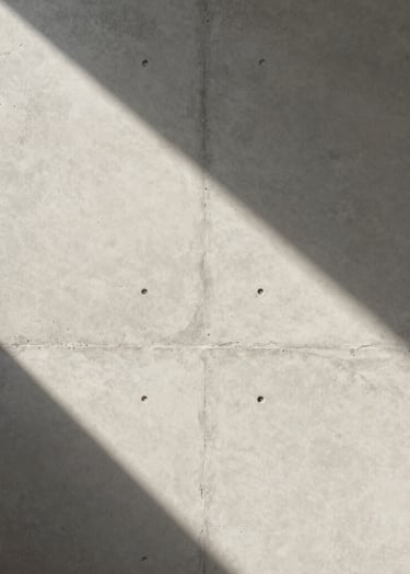 A minimalist detail of a textured concrete wall hit by a sharp sliver of morning light in a Global / Contemporary Art setting. Neutral off-white and soft gray tones. High-end editorial photography style with a focus on geometric shadow.
