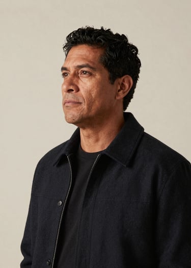 A professional medium-shot photography portrait of a South American man with a thoughtful and soulful expression, wearing a stylish dark jacket, standing against a minimalist cream wall, soft sophisticated lighting, contemporary and authentic.