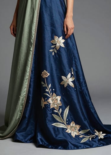 A high-fashion editorial photograph of a silk garment featuring intricate hand-embroidery. The fabric is a deep indigo blue with muted sage green details, draped elegantly in a modern East Asian / South Asian influenced for a global luxury audience studio setting with soft side lighting.