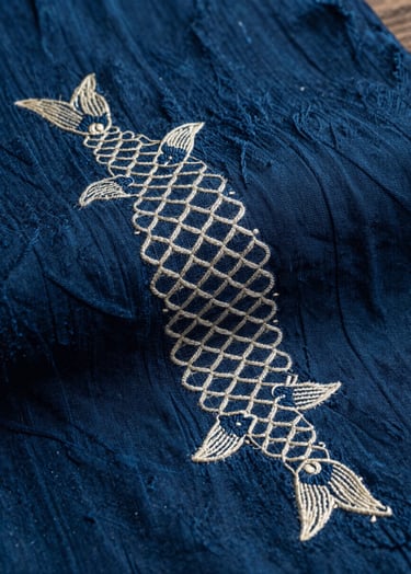 A close-up photograph of hand-woven indigo textile with delicate, ivory silk embroidery of koi fish scales. Cinematic lighting with deep shadows, showcasing the rich texture of the fabric in an East Asian / South Asian influenced for a global luxury audience studio.