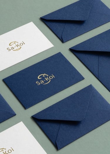 An editorial overhead shot of Sa-Koi visual identity materials, featuring business cards with a minimalist koi logo and deep indigo envelopes on a sage green background, in a East Asian / South Asian influenced for a global luxury audience design office.