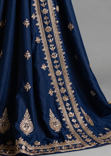 A high-fashion editorial close-up of a textile piece from the Sa-Koi collection. The fabric is a luxurious blend of deep indigo blue silk with intricate Pakistani-style metallic thread embroidery, draped elegantly in an East Asian / South Asian influenced for a global luxury audience studio setting.