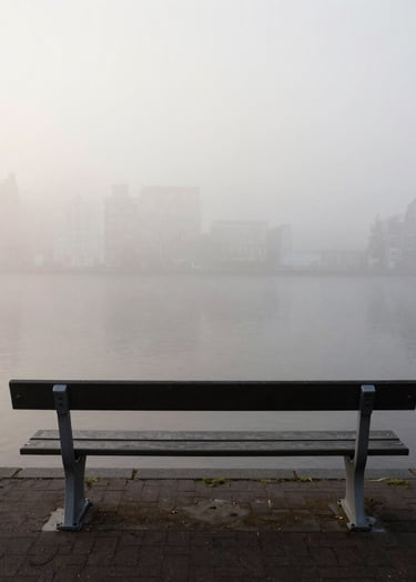 A minimalist, high-contrast photograph of a single empty bench overlooking a foggy canal in Amsterdam. The water is a dark charcoal, the fog is a soft off-white. Quiet, Northern European morning atmosphere, editorial style.