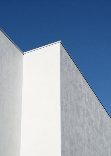 A high-angle architectural photograph of a minimalist white building facade against a clear cerulean blue sky in North America. Sharp, clean shadows create geometric patterns of triangles and rectangles. Bauhaus-inspired composition with a professional, artistic finish.