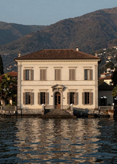 A wide-angle architectural shot of a historic villa on the shores of Lake Como in a North American / European style. The building features clean off-white stone and muted taupe shutters. The water is calm and dark charcoal, reflecting the soft evening light.