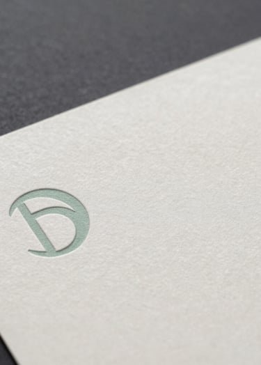 Close-up of premium textured paper with subtle debossed logo details, softly illuminated. The image exudes a professional, minimalist, and strategic aura, reflecting a graphic designer's expertise in visual identities. Color palette incorporates subtle hints of dark navy, slate grey, sage green, and off-white.