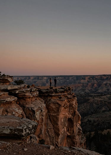 A wide landscape shot of a couple standing on a rugged North American cliff edge at dusk. Cinematic, moody lighting with warm terracotta and charcoal highlights on the horizon. The atmosphere is authentic and vast, shot in a professional lifestyle photography style.
