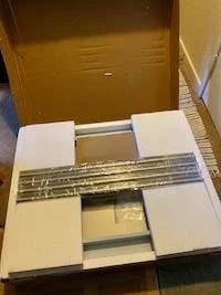 A stainless steel table in its original flat pack packaging with foam protection and assembly parts visible.