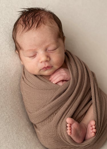 An intimate photography shot of a newborn baby peacefully sleeping, wrapped in a Soft Brown knitted textile. The setting is bright and light with Cream textures, creating a sophisticated and warm atmosphere typical of high-end family photography.