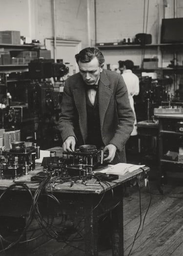 John Logie Baird in a tweed suit and bow tie adjusts a rotating Nipkow disc assembly
