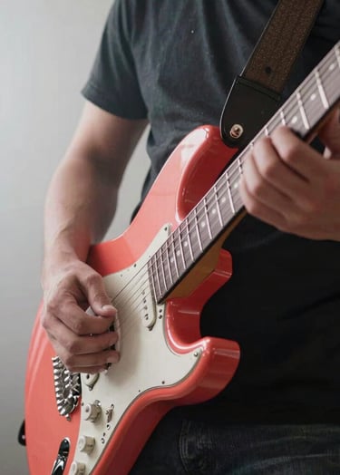 A close-up photograph of a musician's hands playing a Vibrant Coral electric guitar in a bright studio with Light Mist walls. The lighting is energetic and dynamic, highlighting the textures of the instrument in a International / English-speaking setting.
