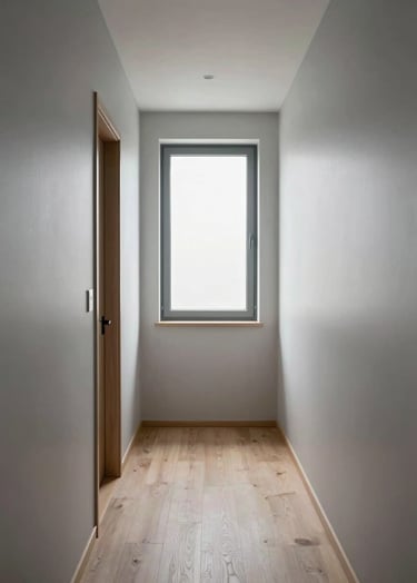 A vertical photograph of a minimalist corridor in a modern Northern European home. The walls are light silver and the floor is pale wood. A single window at the far end provides a bright focal point, creating a sense of depth and tranquility.
