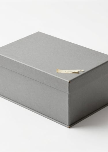 A minimalist, clean shot of a grey archival preservation box sitting on a white surface. A corner of an old, cream-colored manuscript #F8F6F4 is visible. The lighting is soft and even, highlighting the textures of the archival materials.