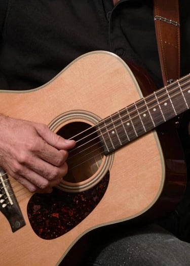 A close-up photograph of a musician's hands gently playing an acoustic guitar in a North American / US music lounge, soft warm lighting, tones of dusty rose brown and deep espresso brown.
