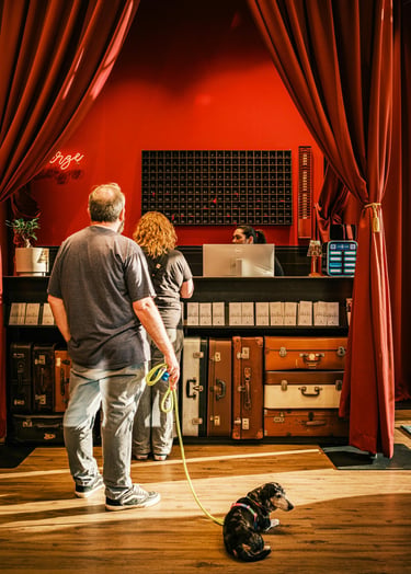 Travelers with a small dog check in at a boutique hotel reception desk with vintage suitcase decor and red curtains.