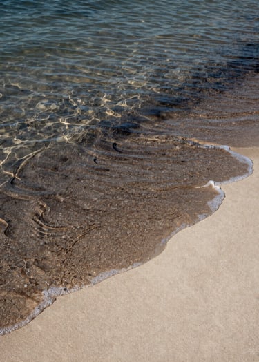 A candid shot of the ocean tide just beginning to wash over a sand pattern. The water is clear, showing the transition between the charcoal-colored wet sand and the light cream-colored dry soft sand.