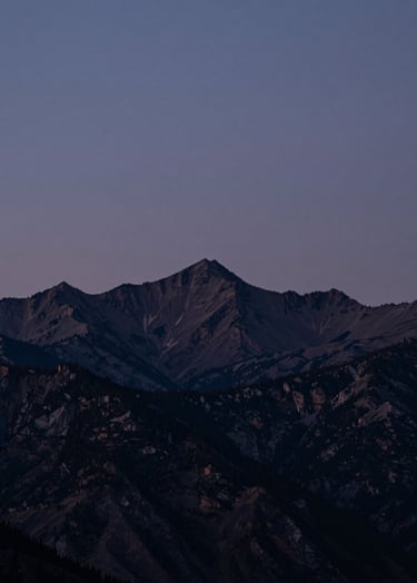 A cinematic wide shot of a North American mountain range at twilight. The sky is a muted indigo, and the silhouettes of the peaks are deep charcoal. The lighting is soft and professional, evoking quiet confidence.