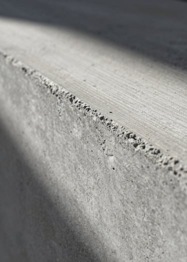 A close-up of textured concrete showing the grain of the wooden formwork. Dramatic side lighting creates a play of light and shadow, emphasizing the material's tactile quality. Minimalist and profound, reflecting the interaction of matter and light.