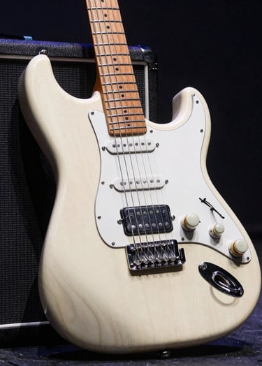 A detailed close-up shot of an electric guitar leaning against a vintage amplifier on a dark stage. The wood grain and chrome hardware are highlighted by soft off-white light against a dark charcoal background. Western European / Dutch concert setting.
