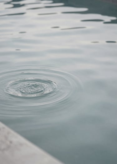 An abstract, minimalist photograph of rippling water in a concrete pool. The tones are muted and neutral, primarily #D0D5DB and #2F363F, reflecting the sky.