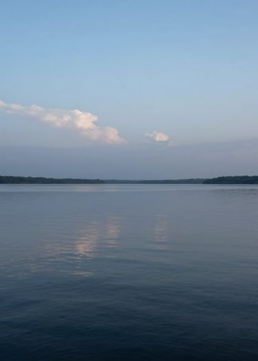 A high-resolution landscape photograph of a calm lake under a pale blue twilight sky. The water reflects the subtle light-grey clouds above. Minimalist and peaceful composition with professional lighting. International / Western nature scene.