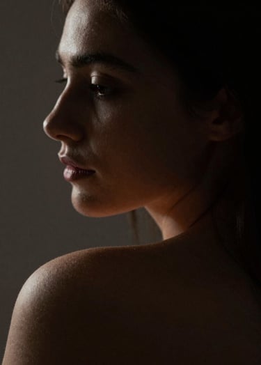 A fine art portrait in extreme chiaroscuro. The light softly catches the curve of a shoulder and the profile of a face, leaving the rest in deep #1F1E1C shadows. Warm, intimate atmosphere with refined textures.