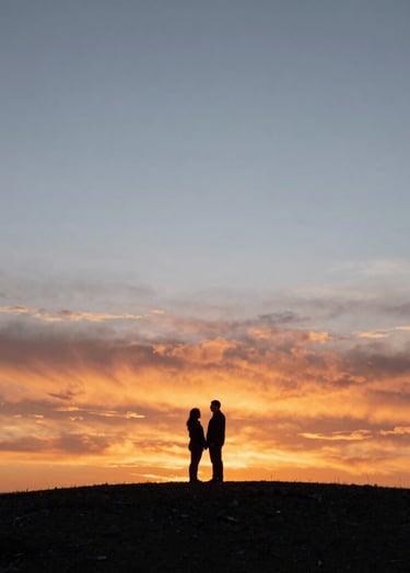 A wide-angle silhouette of a North American / US couple standing on a hill against a vast, cinematic sunset. The sky transitions from a fiery terracotta to a muted light blue. The composition is artistic and focuses on the authentic scale of the landscape and their shared moment.