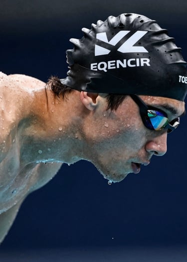 Side profile of a focused athlete in a black swimming cap with white geometric patterns. The lighting is crisp, highlighting water droplets on the skin. The background is a deep dark blue water surface. Western / International setting.