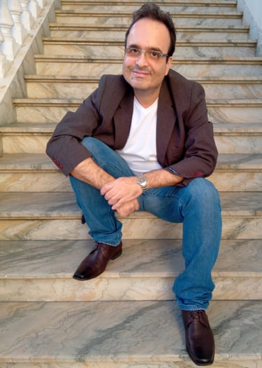 Jornalista Marcelo Dias, sentado à uma escadaria de mármore, de calça jeans, camisa branca e blazer.