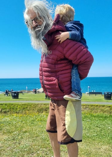 Head surf coach Simon stood with a little boy having a piggy back in green field by the sea.