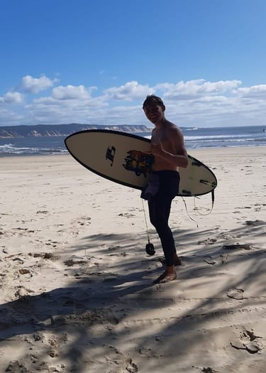 Surf coach Ben standing on a tropical beach holding his surfboard and giving a shaka.