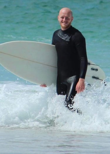 Surf and paddle board coach Lewis (Boris) walking out of a turquiose sea with a surf board