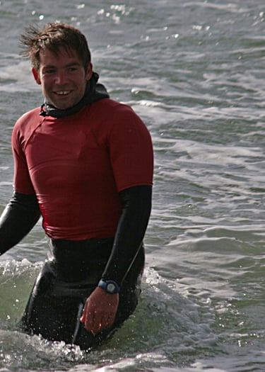 Surf coach Rhys smiling holding the nose of a surfboard in the sea.