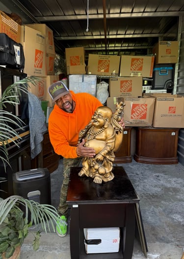 A man in an orange hoodie holds a large golden Buddha statue in a storage unit filled with moving boxes.