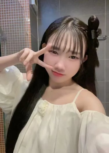 Young woman with long dark hair and bangs wearing a white off-the-shoulder top posing with a peace sign.