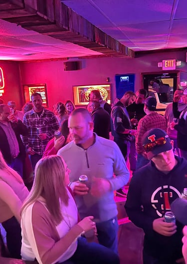 Crowded local bar with patrons enjoying drinks under red and blue neon beer signs.