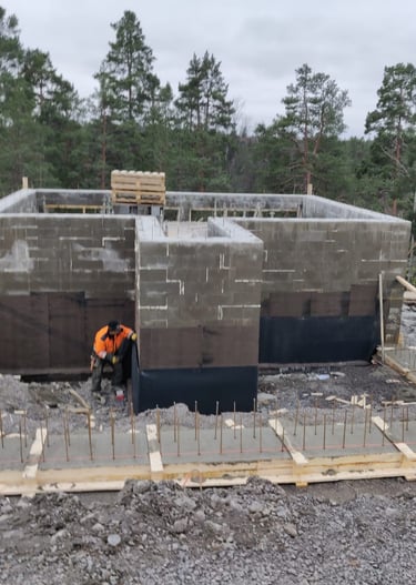 A construction worker builds a concrete block foundation for a new house in a forest setting.