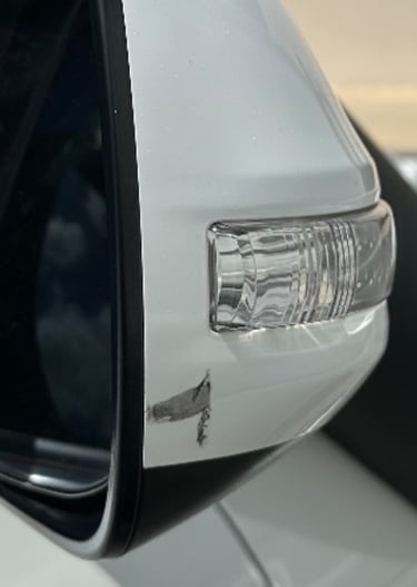 Close-up of a white car side mirror with a scratch and paint damage near the turn signal light.