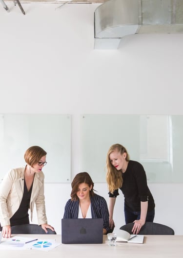 three women standing around a table with laptops