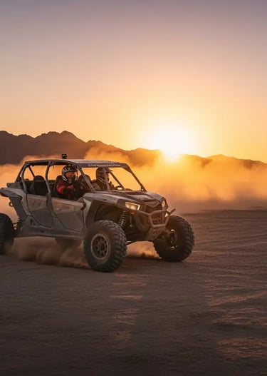 Person driving an off-road ATV through a sandy desert at sunset, kicking up dust.