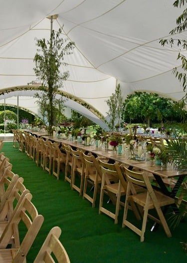 Rustic outdoor wedding reception inside a white marquee tent with long wooden tables and green turf.