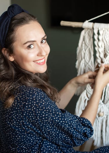 A smiling woman creating a handmade white macrame wall hanging on a wooden easel in her studio.