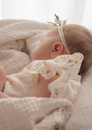 Newborn baby girl sleeping peacefully in a lace outfit and floral headband on a soft white blanket.