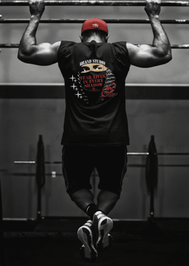 Atleta musculoso con camisa sin mangas negra con estampado gráfico realizando dominadas en un gym.