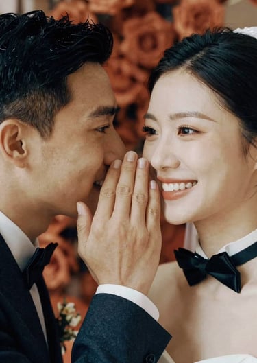 Close-up of a groom whispering to his bride, a genuine smile on her face. Refined elegance in their attire, with terracotta floral accents in the background.