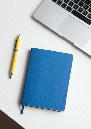 Top-down view of a minimalist desk with a yellow pen, a blue notebook, and a laptop, clean and organized.