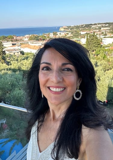 Smiling woman with dark hair on a balcony overlooking a Mediterranean coastal village.