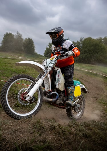 A motocross rider in orange and white gear racing a dirt bike on an off-road track under a cloudy sky.