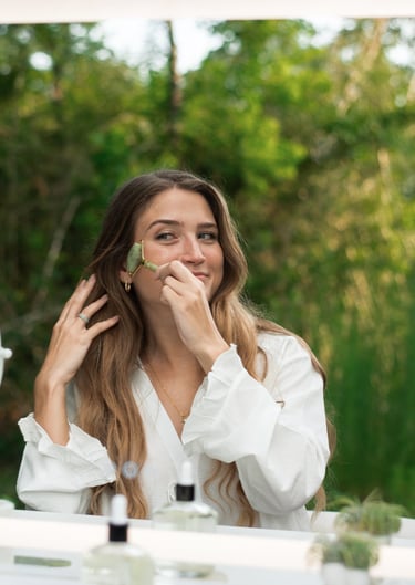 A woman using a green jade facial roller for her skincare routine at a vanity mirror outdoors.